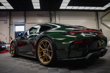 Koyu yeşil bir Porsche 911 GT3, modern bir garajda park edilmiş altın alaşımlı tekerleklerle turluyor. Parlak tavan ışıkları ve yansıtıcı yüzeyler tasarımın altını çizer.