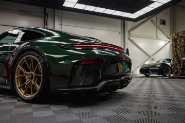 Koyu yeşil bir Porsche 911 GT3 altın tekerlekli turne modern bir garajda park halinde. Arka planda siyah bir Porsche ile arka tasarım ve dört egzoz görünür..