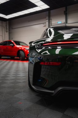 Koyu yeşil Porsche 911 GT3 Touring. Parlak ışıklandırma ve endüstriyel duvarları olan modern bir akort garajında, kareli siyah bir zemine park etmiş..