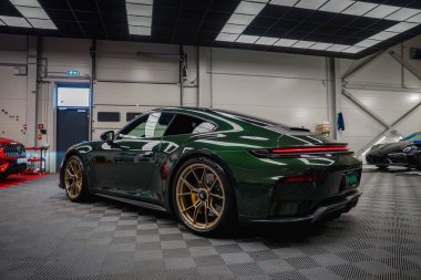 Altın tekerlekleri olan yeşil bir Porsche 911 GT3, beyaz duvarları, siyah kiremitli tavanı ve gri desenli zemini olan modern bir akort garajına park edilmiştir..