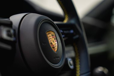 Porsche 911 GT3 Touring 'in direksiyon simidi ayrıntılı olarak gösteriliyor, arka planda Porsche arması, sarı dikişler ve bulanık iç unsurlar yer alıyor..