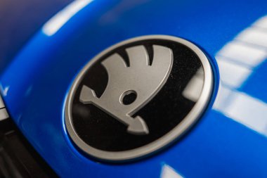 Gümüş kanatlı ok dizaynlı Skoda logosu parlak mavi bir yüzey üzerinde sergilenir, parlak bir kompozisyon içinde yukarıdaki ışıkları yansıtır.