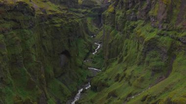 İzlanda 'da sarp yosunlarla kaplı yeşil bir kanyonun havadan görünüşü, belirgin bir şelale, dolambaçlı bir nehir ve arka planda daha küçük şelaleler..