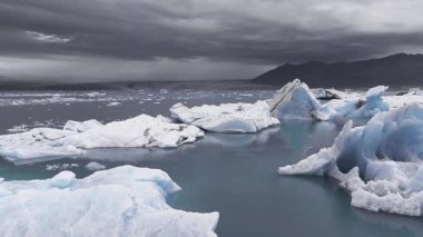 İzlanda 'da, bulutlu bir gökyüzünün altındaki karanlık dağlarla çevrili, sakin sularda sürüklenen buzdağları olan bir buzul gölü..