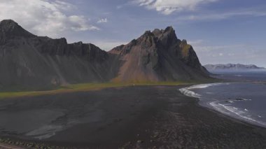 Stoksnes Yarımadası 'nın havasını süpüren Vestrahorn, siyah kumsal, yeşil çimen, sığ su, engebeli uçurumlar ve parçalı bulutlu gökyüzü..