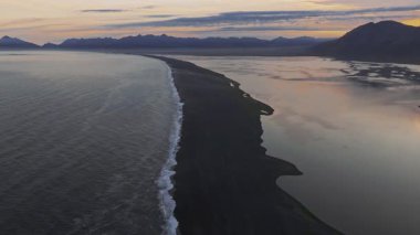 Okyanus dalgaları, yansıtıcı suları ve renkli günbatımı gökyüzünün altındaki uzak dağlarla İzlanda 'da kara bir kumsal manzarası..