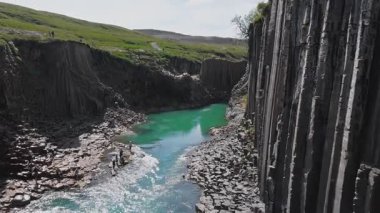Studlagil Kanyonu 'nda bazalt sütunlar, turkuaz bir nehir ve yol boyunca yürüyen ve su kenarında keşif yapan ziyaretçiler bulunur..