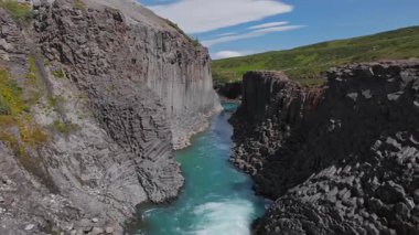 İzlanda 'daki Studlagil Kanyonu' nda bazalt sütunlar, turkuaz bir nehir ve yeşil tepeler bulunur. İnsanlar açık ve güneşli bir günde yürür kenarda.