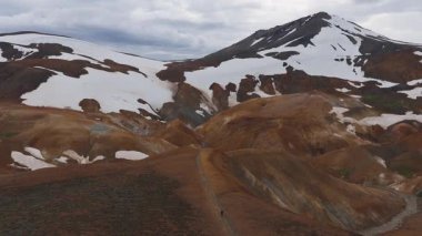 Kar, turuncu ve kahverengi renkli İzlanda dağlarının hava manzarası, kıvrımlı yollar ve hareketli jeotermal oluşumlar..