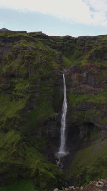 Uzun şelale yosunlu bir uçurumdan aşağı yeşil araziyle çevrili kayalık bir havzaya akar. Arka planda karlı zirveler ve tepeler vardır..