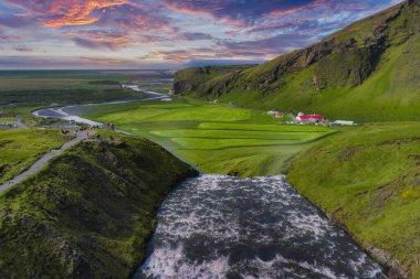 İzlanda 'daki Skogafoss şelalesinin hava manzarası, çağlayan su, yeşil tarlalar, dolambaçlı bir nehir, çiftlik evleri ve renkli bir günbatımı gökyüzü..