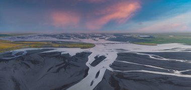 Örgülü buzul nehirleri İzlanda 'da siyah bir kum ovasından akar. Uzak bir köprü ve pembe ve mavi gökyüzünün altında yeşil bir bitki örtüsü vardır..