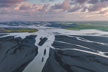 İzlanda 'da gün batımında pembe ve mor bir gökyüzü altında volkanik kumlar, bir köprü ve yeşil tarlalarla örülmüş nehir sisteminin hava görüntüsü..