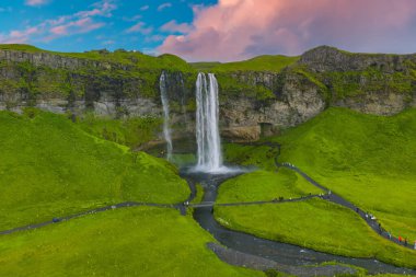 İzlanda 'da Seljalandsfoss şelalesi yeşil tepelerle çevrili yüksek bir uçurumdan dökülüyor, dolambaçlı bir yol ve pembe ve mavi gökyüzünün altında sakin bir dere..