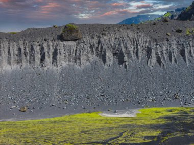 İzlanda 'da sarp bir volkanik uçurum aşınmış dokuları ve parlak yeşil yosunlarıyla karanlık toprakla çelişiyor. Pembe ve turuncu gökyüzü ve uzak dağlar sahneyi tamamlıyor..