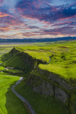 İzlanda 'daki yemyeşil uçurumların havadan görünüşü küçük bir şelale, dolambaçlı bir yol, dağınık binalar, pembe ve turuncu renkli bir günbatımı gökyüzü..