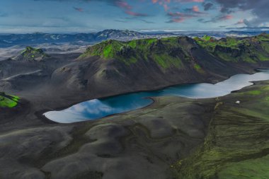 Yansıtıcı gölü, yosun kaplı tepeleri, uzak dağları ve dramatik pembe ve mavi gökyüzü olan İzlanda 'daki volkanik bir arazinin havadan görünüşü..