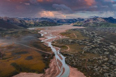 İzlanda 'da gün batımında karlı dağlarla kaplı, yosunlu tepeler, kayalık çıkıntılar ve topraksı arazi boyunca kıvrılan örülmüş nehir sisteminin havadan görüntüsü..