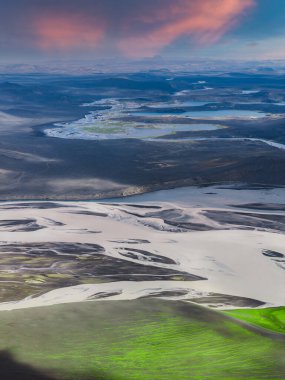 Yeşil yosun, volkanik arazi, uzak göller ve pembe ve turuncu renkli gökyüzü ile İzlanda 'da örülmüş bir nehir sisteminin hava görüntüsü..