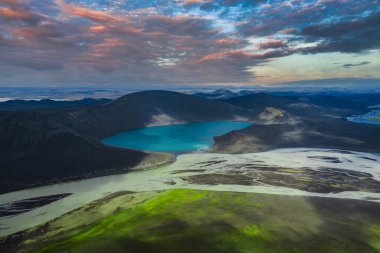 Turkuaz göl koyu volkanik arazi, soluk nehir kanalları ve yeşil yosun ile çevrili parlak turuncu, pembe ve mavi günbatımı gökyüzü altında.
