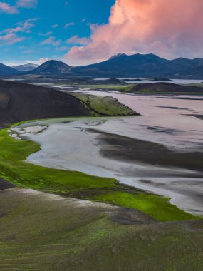 Yeşil yosun ve volkanik toprakla çevrili, pembe ve mavi gökyüzünün altında engebeli dağlarla çevrili kıvrımlı bir nehrin hava manzarası..