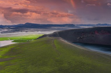 İzlanda 'da volkanik kratere giden bir yolu olan yeşil bir tepenin havadan görünüşü, tabanında bir göl, uzak ovalar, nehirler, dağlar ve canlı bir günbatımı altında bir gökkuşağı..