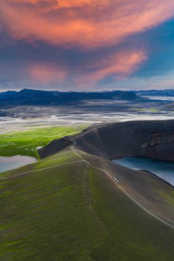 İzlanda 'daki bir volkanik kraterin hava görüntüsü, kısmen suyla dolu, çevresi yeşil yosun, karanlık toprak, düzlükler ve günbatımında uzak dağlarla çevrili..