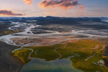 Örülmüş nehirler yosunlu yeşil ve sarı arazi boyunca volkanik topraklarla akar, engebeli dağlara, buzullara ve renkli bir günbatımı gökyüzüne karşı kurulur..
