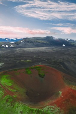 İzlanda 'daki volkanik bir elma kraterinin havadan görünüşü, canlı kırmızı ve yeşil renkler, koyu arazi, karla kaplı dağlar ve renkli bir gökyüzü..