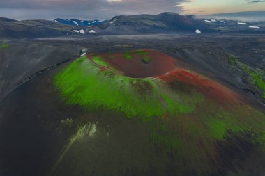 Yeşil yosun, kırmızı toprak ve karanlık arazisi olan İzlanda 'daki volkanik bir elma kraterinin havadan görünüşü. Karla kaplı zirveler uzak arkaplanda görülebilir.