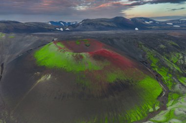 Yeşil yosun, kırmızı toprak ve karanlık arazisi olan İzlanda 'daki volkanik bir elma kraterinin havadan görünüşü. Karlı dağlar ve pastel gökyüzü güneşin doğuşunu veya batışını akla getiriyor..