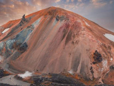 Landmannalaugar, İzlanda 'daki canlı rhyolite dağı, turuncu, kırmızı ve sarı renkler, yamaçlardaki yürüyüşçüler, jeotermal buhar ve sıcak tonlu gökyüzü..