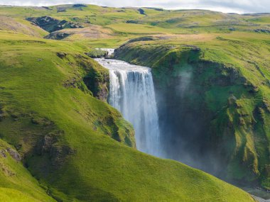 Geniş çağlayan Skogafoss şelalesi dalgalı yeşil tepeler ve sisli spreylerle çevrili sarp bir uçurumdan aşağı düşer..