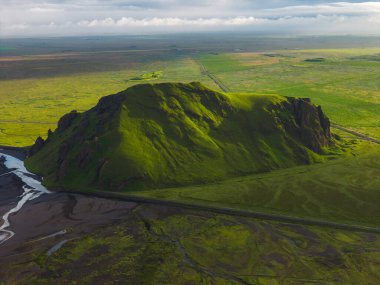 Düzlüklerle, kıvrımlı derelerle ve İzlanda 'da sulak alanlarla çevrili yeşil bir tepe. Bulutlu, güneşli bir gökyüzünün altında bir yol var..