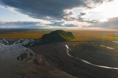 Issız yeşil bir tepe İzlanda 'nın düz bir volkanik manzarasından yükseliyor, etrafı örülmüş nehirlerle çevrili ve dramatik bulutların altında altın gün batımı ışığıyla aydınlatılıyor..