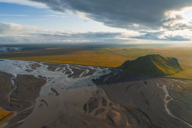 Örgülü nehir kanalları İzlanda 'da düz bir ovada akıyor, yeşil bir tepe hızla yükseliyor. Altın ışık ve dramatik bulutlar sahneyi güçlendirir.