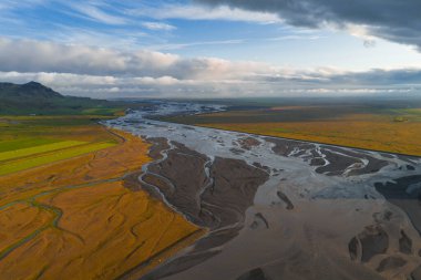 İzlanda 'da örülmüş bir nehir sisteminin havadan görünüşü, yeşil ve altın tarlaları, karanlık nehir yatakları, uzak dağlar ve parçalı bulutlu bir gökyüzü..