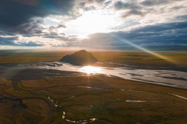 Issız bir kaya oluşumunun yer aldığı geniş İzlanda arazisinin havadan görünüşü, güneş ışığını yansıtan dolambaçlı nehirler ve İzlanda 'daki bulutların arasından yükselen sıcak ışık..