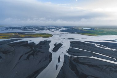 Gümüşi kanallar, karanlık volkanik kumlar, bir köprü, yeşil bitki örtüsü ve İzlanda 'da bulutlu bir gökyüzü ile örülmüş bir nehir sisteminin hava görüntüsü..