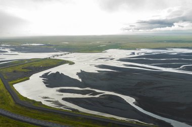 Örülmüş buzul nehirleri, yeşil tarlalar ve İzlanda 'da parçalı bulutlu bir gökyüzü ile çevrili bir köprü geçidi ile siyah kum düzlüklerinde akar..