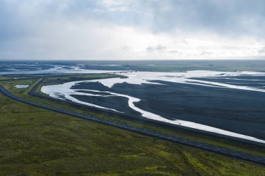 İzlanda 'da, yosunlu araziyle çevrili karanlık volkanik kumları kesen örülmüş bir nehrin havadan görünüşü. Asfalt bir yol ve köprü bulutlu bir gökyüzünün altında görünür..