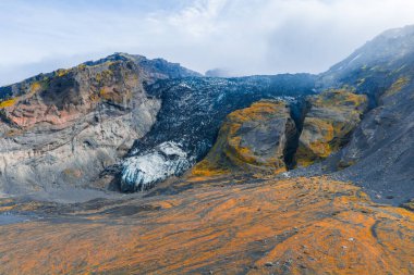 Sarp, renkli kaya oluşumlarıyla dramatik bir İzlanda kanyonu ve bulutlu bir gökyüzünün altında kayalıkların arasına yerleşmiş bir buzulla çarpıcı bir manzara yaratıyor..