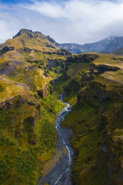 İzlanda 'daki Mulagljufur Kanyonu' nda dik yosun kaplı uçurumlar, dolambaçlı bir nehir, canlı yeşil bitki örtüsü ve bulutların altındaki uzak kar tepeleri bulunur..