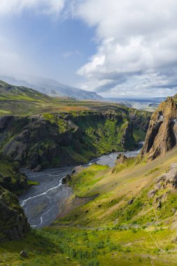 İzlanda 'daki Mulagljufur Kanyonu' nun hava manzarasında, bulutlu bir gökyüzü altında dramatik uçurumlar, dolambaçlı bir nehir, yemyeşil bitki örtüsü ve buzul özellikleri yer alıyor..