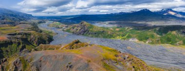 Yeşil ve kahverengi tepeleri, volkanik kaya oluşumları ve İzlanda 'daki uzak kar tepeli dağları olan bir vadiden geçen örülmüş nehirlerin havadan görüntüsü..