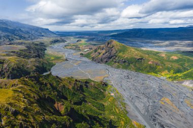 Yeşil tepeler, kayalık tepeler, volkanik topraklar ve bulutlu bir gökyüzünün altındaki uzak dağlarla geniş bir vadiden geçen İzlanda 'da örülmüş bir nehrin havadan görünüşü..