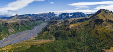 İzlanda 'da yeşil bir vadiden süzülen bir buzul nehrinin hava manzarası. Etrafı dramatik dağlar ve parçalı bulutlu bir gökyüzünün altındaki kar tepeleriyle çevrili..