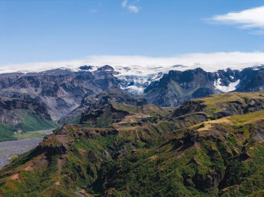 Yeşil tepeler, kayalık vadiler ve İzlanda 'da dağınık bulutlarla dolu mavi bir gökyüzünün altındaki sivri dağlarla çevrili devasa bir buzul manzarası..