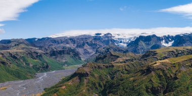 İzlanda 'da dolambaçlı bir nehir, kayalıklarla çevrili yemyeşil bir vadiden akar. Arka planda açık mavi bir gökyüzünün altında devasa bir buzul bulunur..