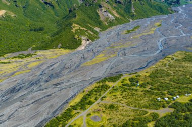 Bereketli yeşil vadide örülmüş bir nehir sisteminin hava manzarası engebeli tepeler, karanlık çökeltiler ve İzlanda 'da çadırları ve araçları olan bir kamp alanı..